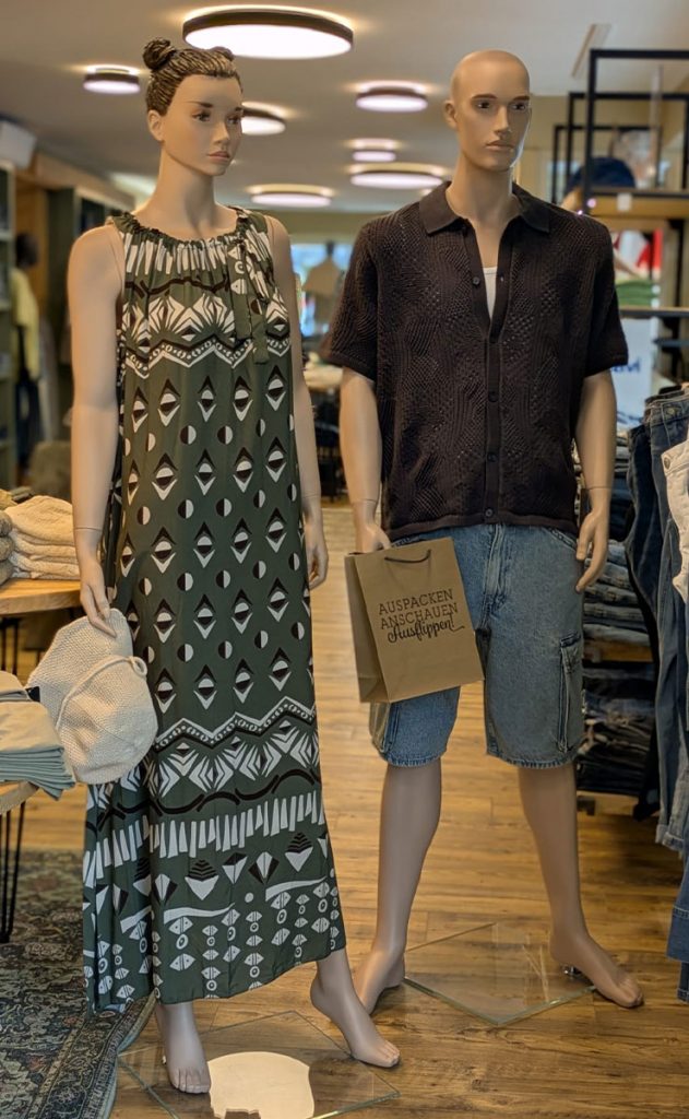 Sommermode 2026 im Jeans House Olching mit gemustertem Maxikleid, schwarzem Kurzarmhemd und Denim-Shorts für Damen und Herren.