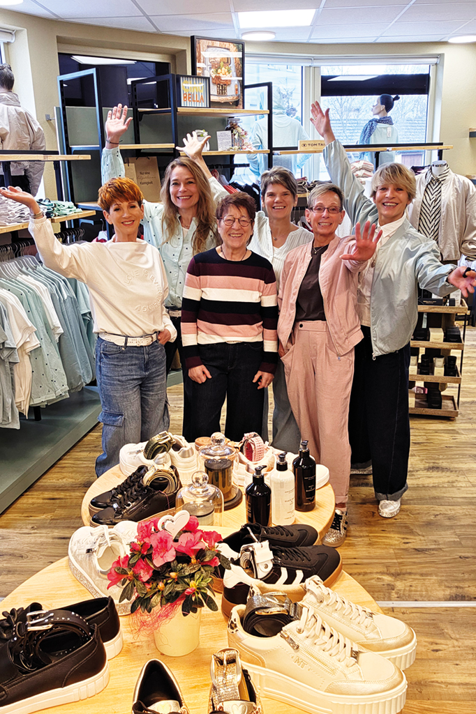 Sechs fr&ouml;hliche Mitarbeiterinnen des JEANS HOUSE Olching begr&uuml;&szlig;en l&auml;chelnd die Kundschaft im Gesch&auml;ft. Im Vordergrund stehen modische Sneaker, Accessoires und Duftprodukte auf einem Holztisch, w&auml;hrend im Hintergrund helle Sommermode und Jeanskollektionen pr&auml;sentiert werden. Die freundliche Atmosph&auml;re vermittelt pers&ouml;nliche Beratung und Freude an Mode.