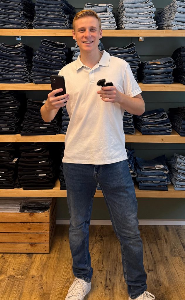 Ludwig, Social-Media-Experte im JEANS HOUSE Olching, steht mit Smartphone und Mikrofon vor einem Regal voller Jeans und erstellt kreative Inhalte für die Social-Media-Kanäle des Modegeschäfts.